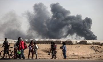 Plumes of grey smoke ascend from rows of tents as a group of men women and children walk past in the foreground