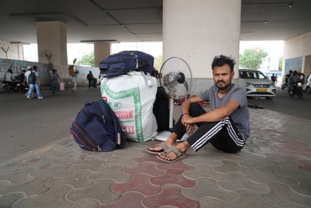 A man sits on the floor next to several large bags and a fan.