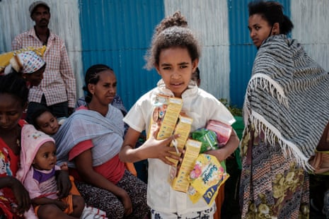 A girl holds food items and looks at the camera as women look on behind her.