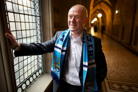 Sir Richard Leese smiles, wearing a Manchester City scarf, in Manchester town hall