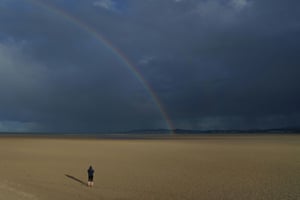 A person looks at a rainbow and the heavy rain in the distance, while walking on an empty beach in sunshine in Dublin