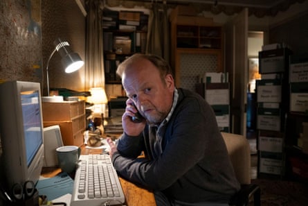 Toby Jones on the phone at a desk and keyboard with a lamp shining down in the dark
