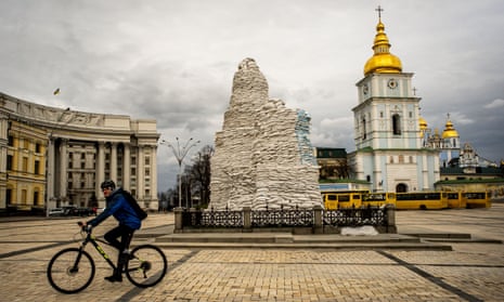 Man cycles past statue, which is entirely covered by high pile of sandbags. Church tower with golden dome and blue and white plaster is seen to the right