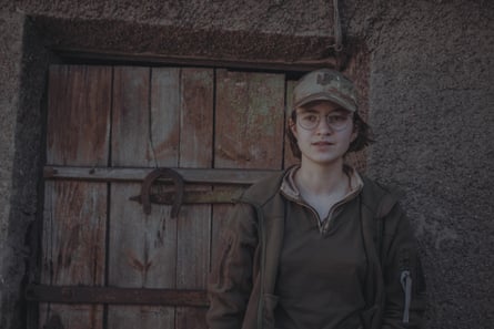 A young woman wearing a camouflage-patterned cap, khaki clothes and glasses stands in front of a wooden door