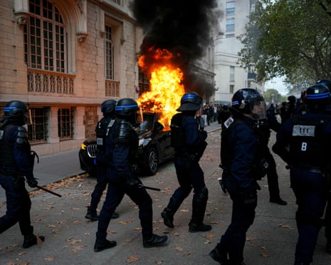French police officers in riot gear walk past a burning vehicle during a demonstration of high-school students in Paris.