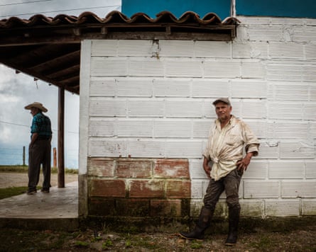 An older man in dirty work clothes and rubber boots leans against a wall while around the corner another man in a cowboy hat stands on a veranda