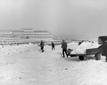 Teams of men clear deep, thick snow from the track at Cheltenham racecourse, shovelling it on to a cart; the grandstands are in the background