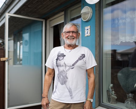 Kári Böðvarsson stands smiling outside his home. He is 79 with a white beard and white hair, and wears a white T-shirt with an image of what appears to be a naked man wearing only sunglasses and a cowboy hat and playing a strategically placed bass guitar. There are lace curtains in the window of the house.