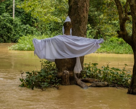 A brown river in spate flowing past a crude wooden sculpture of a figure draped in a white cloth