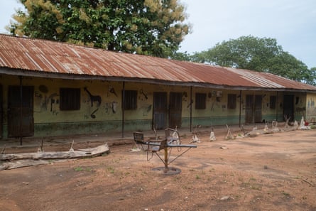 A single story school house, painted yellow and green with an illustrated alphabet on the walls. A broken roundabout is in the dusty yard.