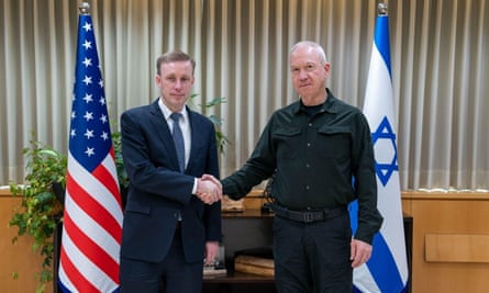 Jake Sullivan shakes hands with Yoav Gallant: they are both facing the camera for a formal photo and are standing in front of their respective national flags in a conference-style room with wooden units and white curtains.