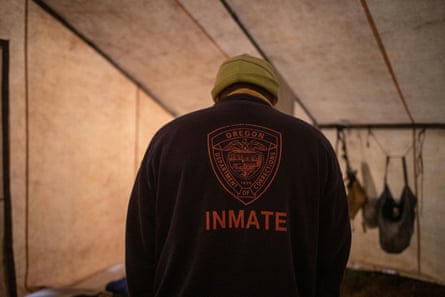 A person stands in a tent with their head bowed. On the back of the black sweatshirt they are wearing are the words ‘Inmate’ and ‘Oregon department of corrections’.