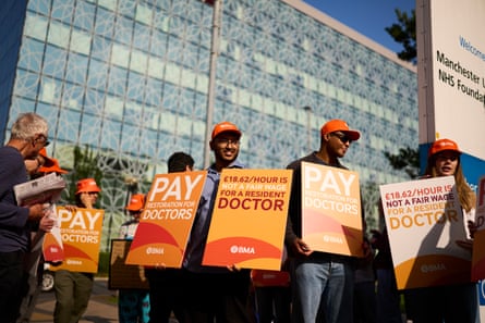 two young men and a woman in orange hats holding placards stand in front of others; the tall, glass-fronted hospital building is behind them
