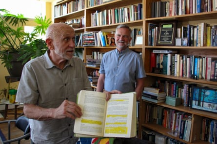 A man shows highlighted lines from a book in his home library while another man looks on