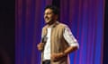 Ahir Shah, a thirty-something man, wearing glasses and a white shirt with a light brown sleeveless jacket over it, stands at a microphone looking upwards, with a blue-and-red-lit curtain behind him.