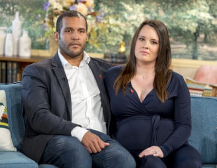 A man of colour sitting on a sofa wearing a suit and a woman in a black outfit, with long dark hair, both sitting on a blue sofa with greenery behind them