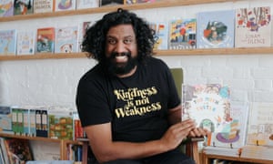 A man, smiling cheerfully, sits in front of a wall of children's books, holding a picture book, as if about to read