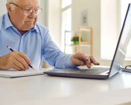 Senior man in glasses using laptop and taking notes in notebook sitting at desk at home