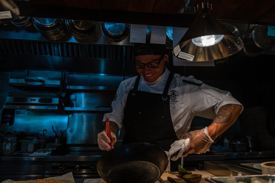 A man in a chef’s apron holds a cast iron pan in one hand as he cooks at an industrial stove.