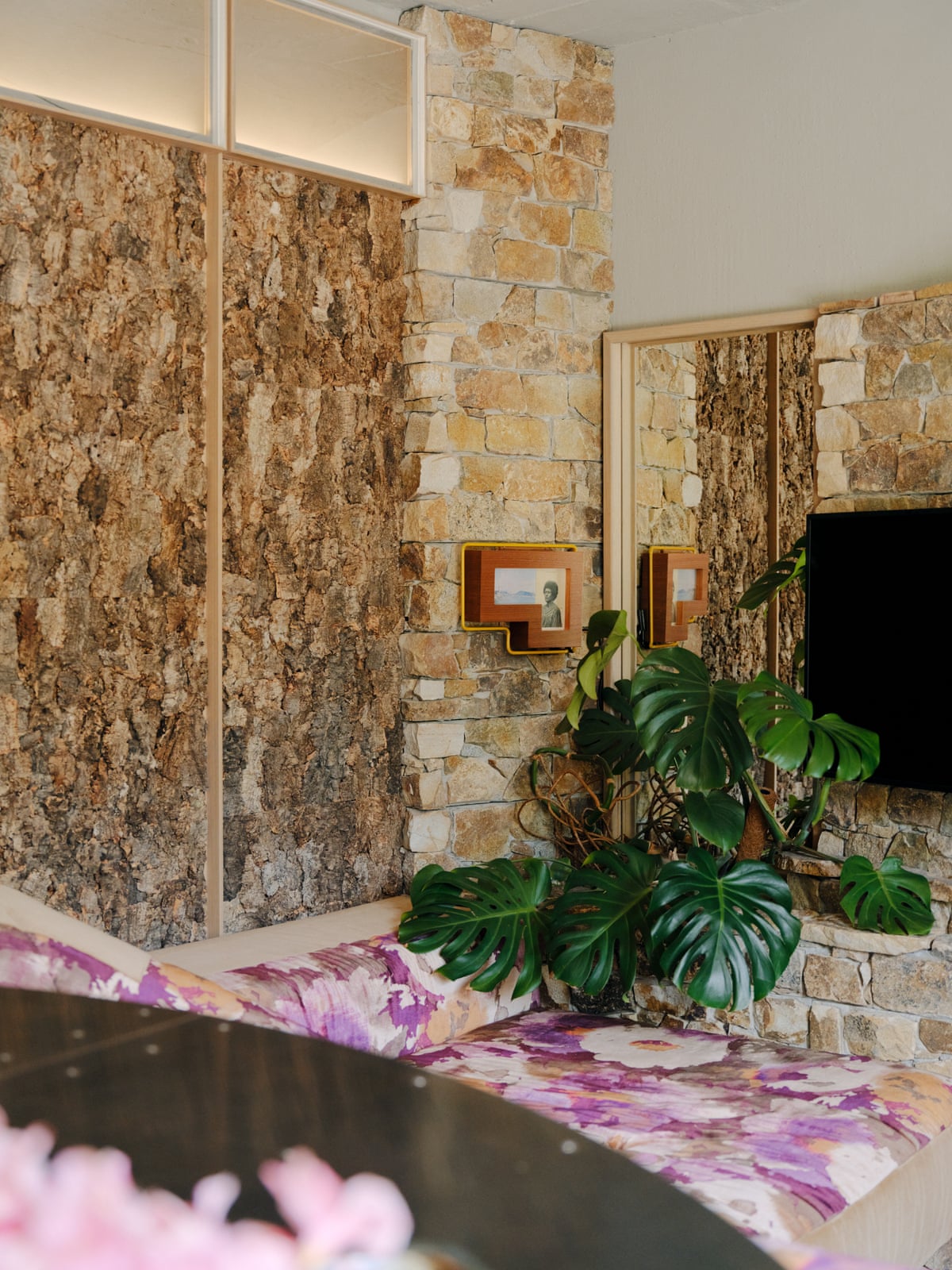 A stone and cork-walled apartment interior with a monstera plant, television, and floral seating