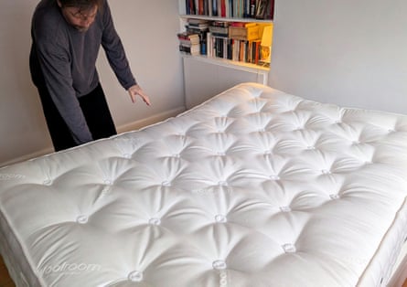 Man in a long-sleeved shirt bends down to touch the edge of a bare mattress with books on shelves to his left