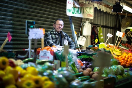 A market trader stands behind a stall of colourful fruit and vegetables.