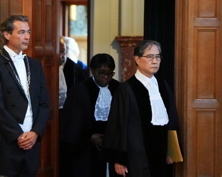 Yūji Iwasawa arrives at the court: he walks through a wood-panelled doorway holding a folder of documents. He wears a white shirt with judicial ruff and a black jacket.