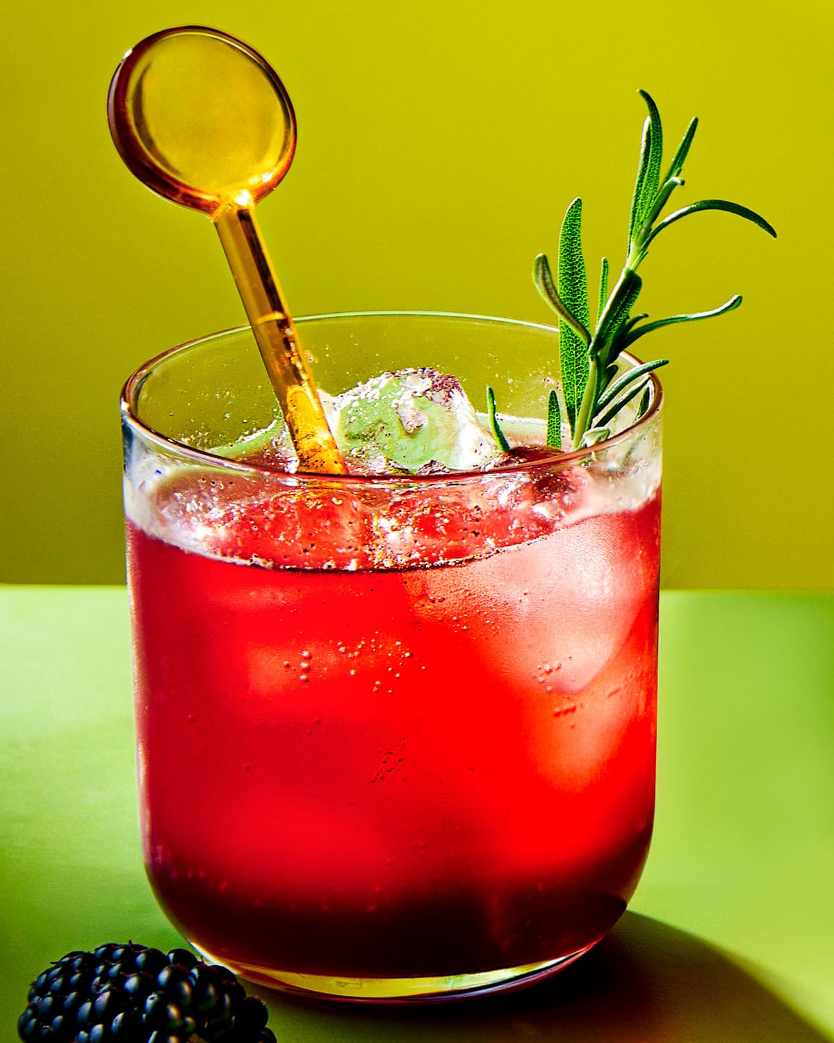 A bramble and ginger shrub sits redly, in a cocktail tumbler, upon a green table, with an orange transparent plastic cocktail stick in it, and a fetching spring of rosemary. And a blackberry lies at its base.