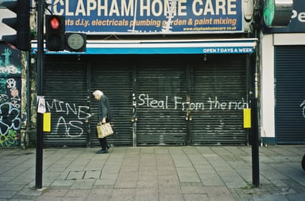 Colour photograph of an elderly person walking along the street past a shuttered shop front that has ‘Steal from the rich” spray-painted in front.