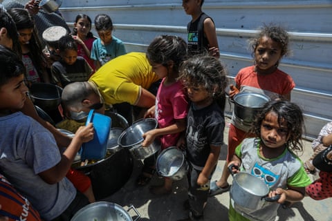Young girls and boys stand against a metal fence holding pans and bowls. qhxidiqxkiqduinv