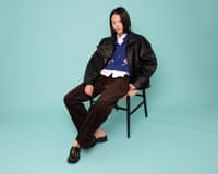 A female model in a black leather jacket, blue cardigan and brown trousers sits on a chair against a turquoise background