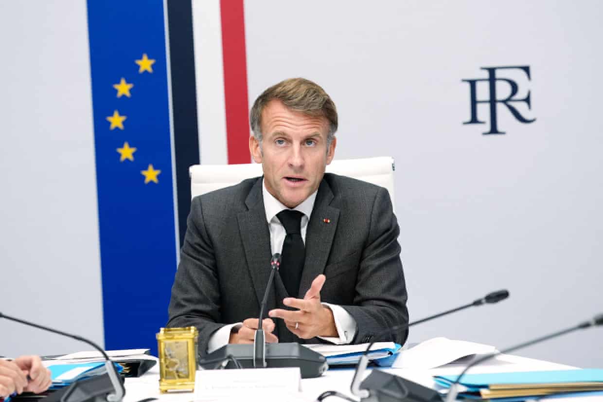 French president Emmanuel Macron sitting at table in front of vertical banners of the EU and French flags.