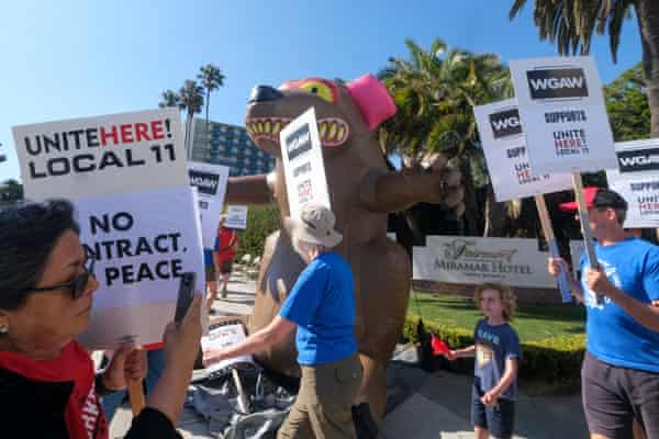 Workers picket in from of an inflatable rat outside a hotel under palm trees.