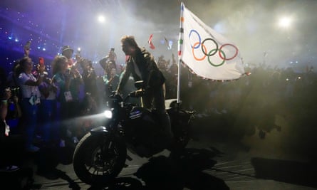 Tom Cruise drives out of the Stade de France on a motorbike with the Olympic flag