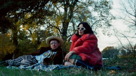A man lying and a woman in a red coat sitting on a blanket on grass near trees
