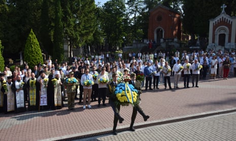 Two Ukrainian soldiers carry a wreath of blue and yellow flowers in front of a crowd of Orthodox priests and others holding bouquets