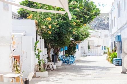White houses on a narrow white paved street with a tree and blue outdoor chairs
