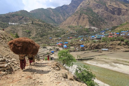 People walk down a steep rocky pathway, carrying huge loads of a dark brown crop on their heads, towards a green valley with scattered houses with colourful roofs, surrounded by mountains.
