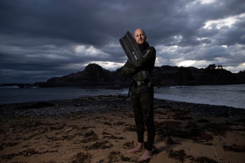 A freediver in a wet suit holding a swimming fin, posing on a beach at dawn.