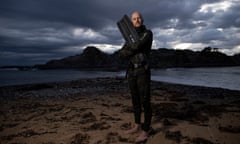A free-diver in a wet suit holding a swimming fin, posing on a beach at dawn.