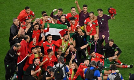 Um grupo de jogadores de camisa vermelha se reúne para uma foto em campo, gesticulando em comemoração.  Vários jogadores levantam a bandeira da Palestina no meio do grupo