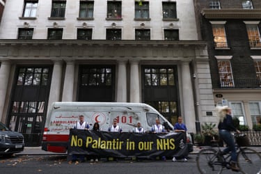 A van made to look like a heavily damaged Gaza ambulance is parked outside Palantir’s headquarters. A line of protesters stand in front of the ambulance holding a sign that says ‘No Palantir in our NHS’