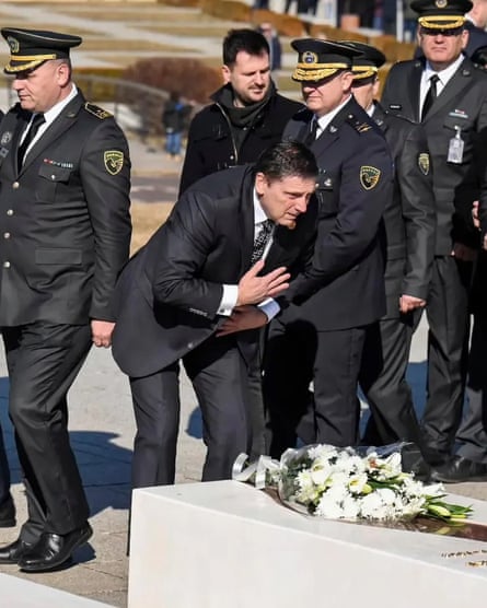 Politician bows before grave with white flowers on it as men in military uniforms walk behind him