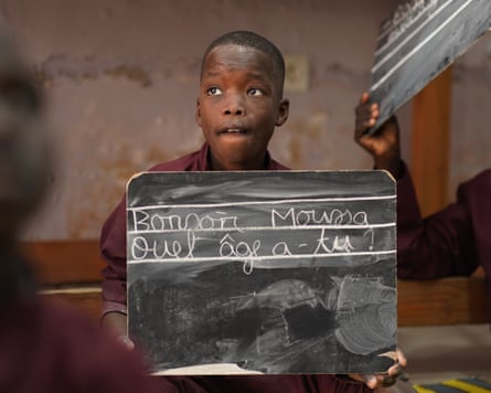 A young boy with a hopeful expression holds up a small blackboard with words in French written in chalk