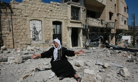 An elderly woman holds her hands apart as she sits in front of damaged houses