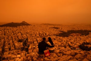 A couple sits on a hill looking out over Athens under an orange sky