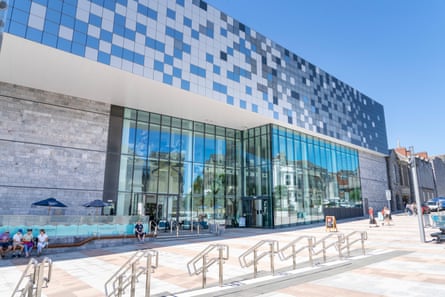 People sit outside a large glass and mosaic-tiled building on a sunny day