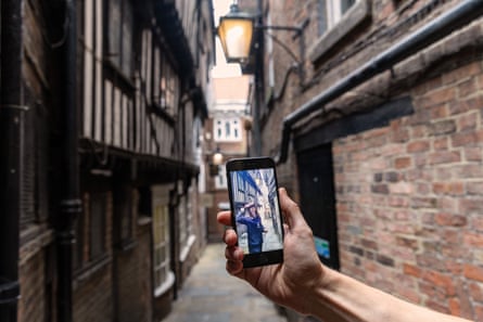 A female character appears on a phone screen as the phone is held up in front of a narrow alleyway of ancient half-timbered buildings.