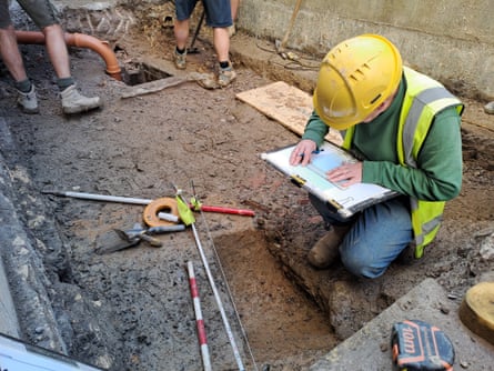A man in a yellow hard hat kneeling in an excavated ditch with measuring tape on the ground records measurements on a clipboard