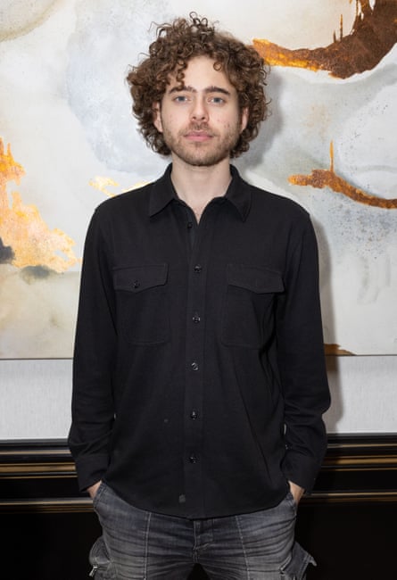 A man with curly hair poses in a button-down shirt and jeans